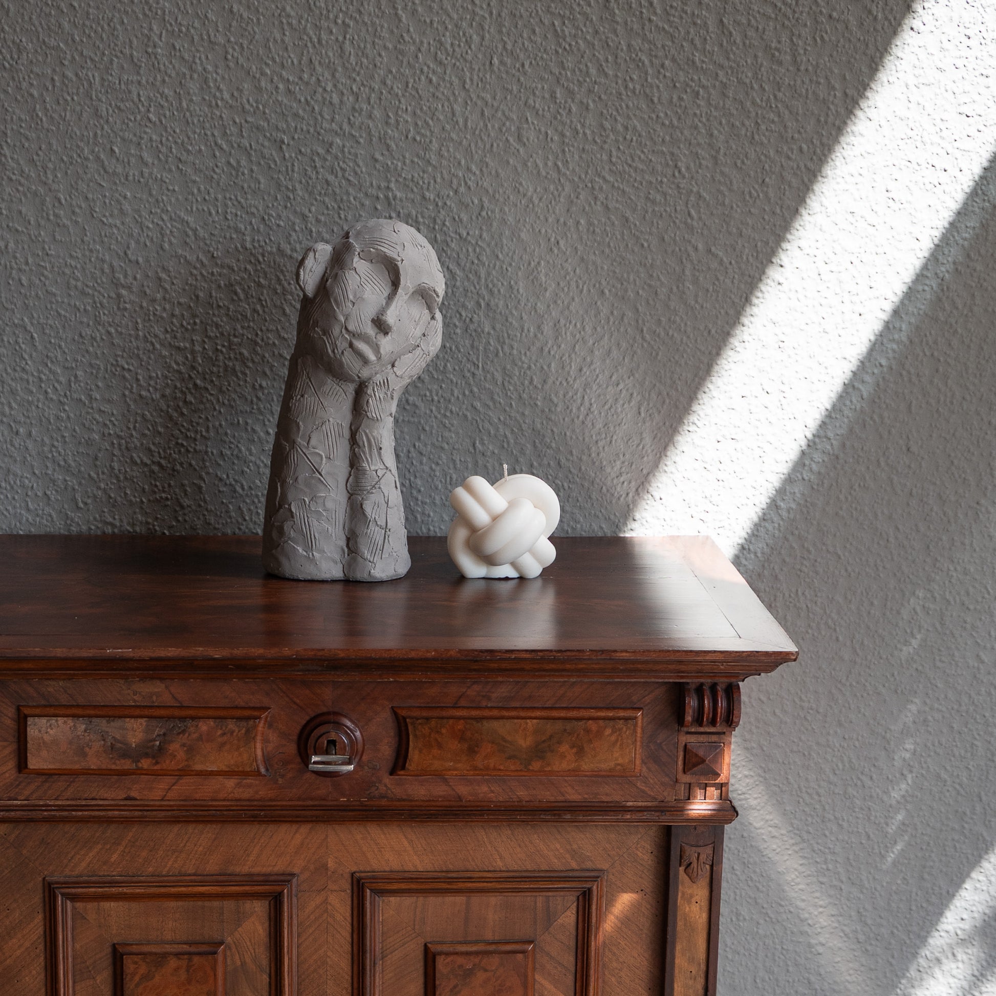 Skulpturale Designkerze im skandinavischen Stil. Handgfertigt in Hamburg aus 100% Rapswachs. Minimalistisch, ästhetisch, zeitlos.  Knoten. Dekoriert auf barocker Kommode im Sonnenlichtspiel neben Dekofigur.