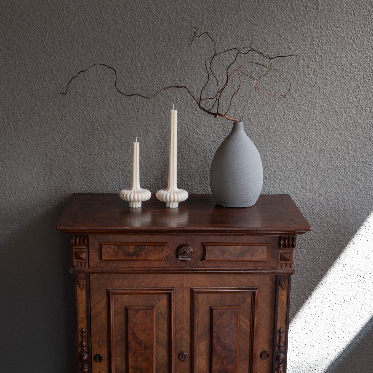 Zweier Set Skæva Kerze in natürlichem Cremeton – skulpturale Designkerzen aus Rapswachs, handgefertigt in Hamburg, 100 % pflanzlich & nachhaltig. Studio Z. Mit plissierter Struktur, markantem Fuß und skulpturaler Form. Dekoriert nebeneinander in zwei Größen auf barockem Tisch neben grauer vase mit drahtigem Ast vor grauer Wand.