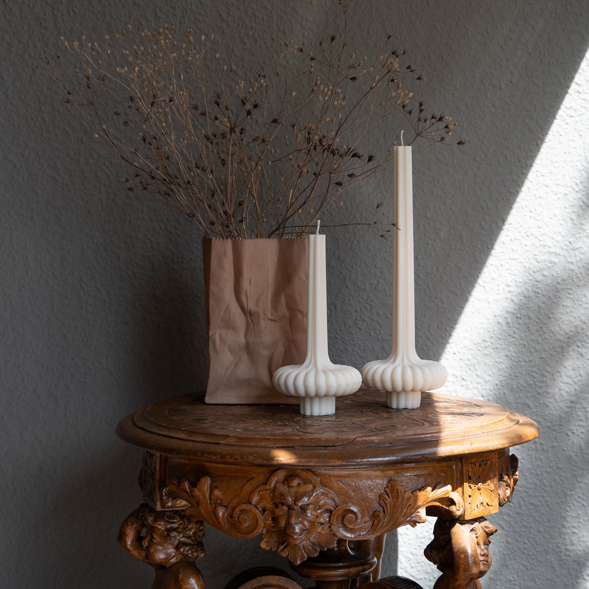 Zweier Set Skæva Kerze in natürlichem Cremeton – skulpturale Designkerzen aus Rapswachs, handgefertigt in Hamburg, 100 % pflanzlich & nachhaltig. Studio Z. Mit plissierter Struktur, markantem Fuß und skulpturaler Form. Dekoriert nebeneinander in zwei Größen auf barockem Tisch vor grauer Wand neben Vase mit Blumenzweigen und Schattenspiel durch Sonnenlicht.