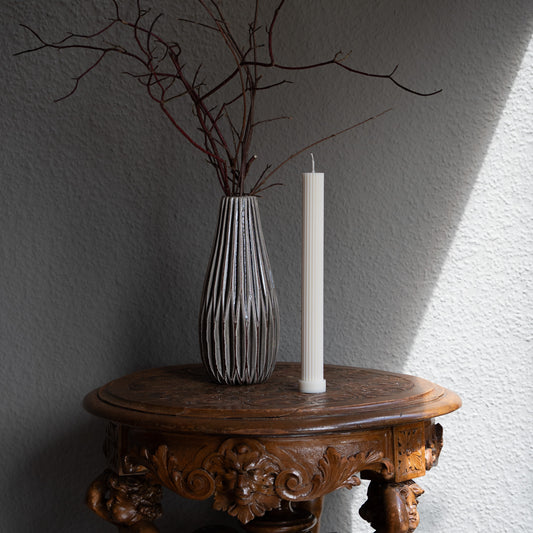 Kerze Lys, geradlininge Stumpenkerze in natürlichem Cremeton – geriffelte längliche Designkerzen, Rapswachs, handgefertigt in Hamburg, 100 % pflanzlich & nachhaltig. Studio Z. Dekoriert auf barockem Tisch vor grauem  Hintergrund neben geriffelter Vase mit Ästen.
