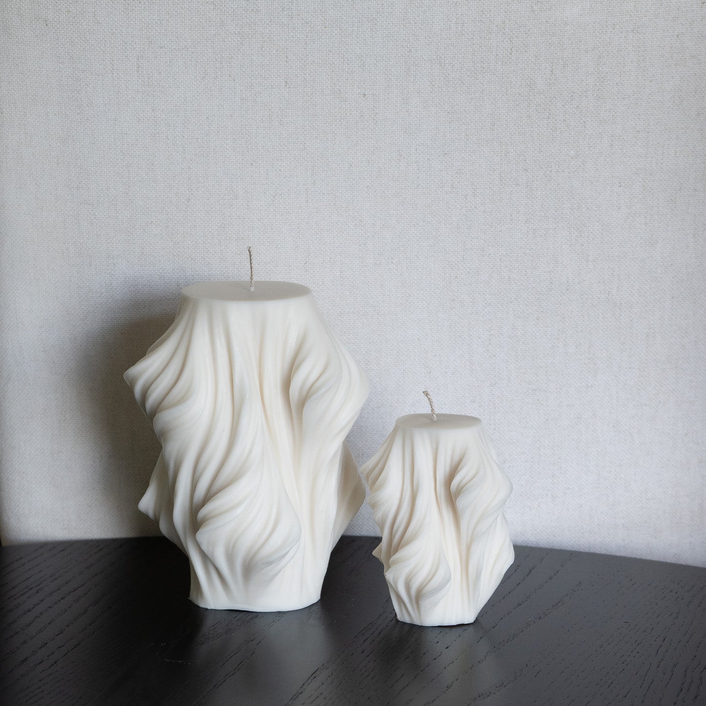 Skulpturale Kerze in Naturfarbe. Handgefertigt in Hamburg – Statement-Dekor im dänischen Design. 100 % natürliche, nachhaltige Materialien. Jedes Stück ein Unikat. Schleier Optik. Set aus zwei Kerzen, zwei Größen. Dekoriert auf schwarzem Tisch vor Leinenstuhl.