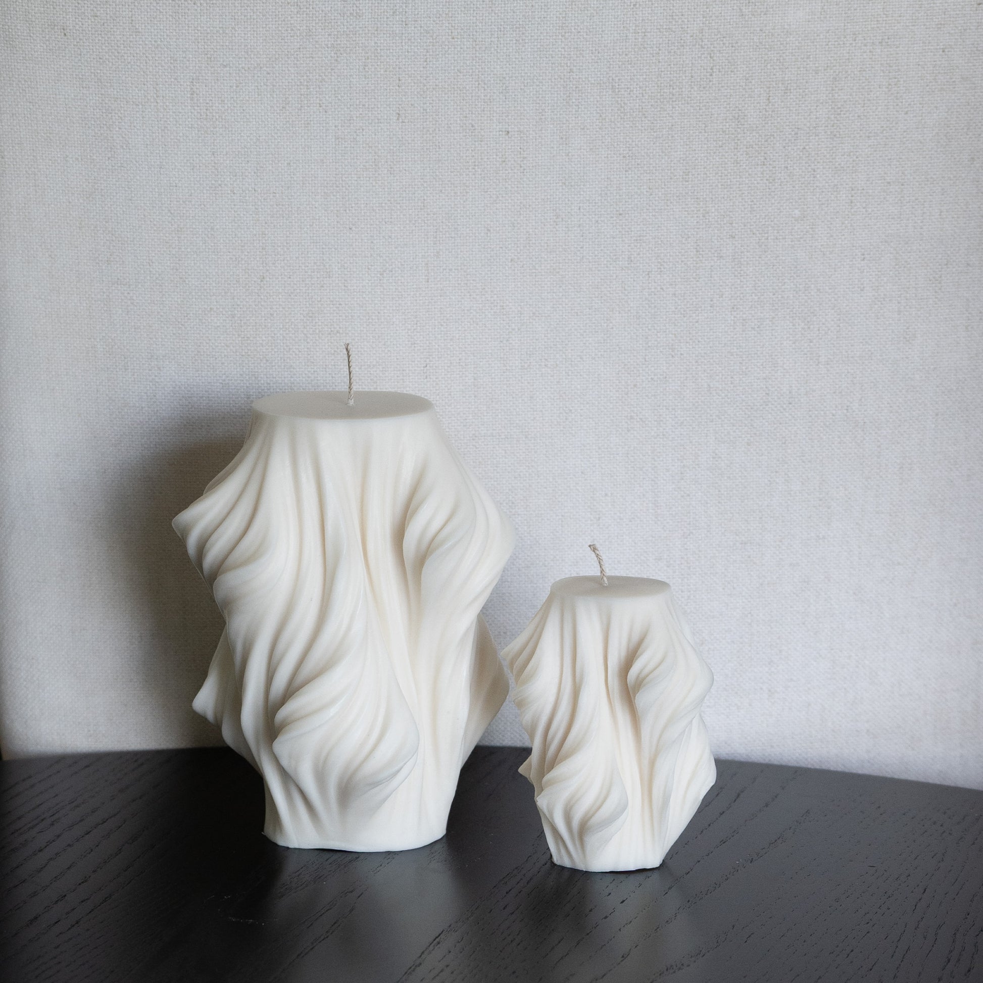 Skulpturale Kerze in Naturfarbe. Handgefertigt in Hamburg – Statement-Dekor im dänischen Design. 100 % natürliche, nachhaltige Materialien. Jedes Stück ein Unikat. Schleier Optik. Set aus zwei Kerzen, zwei Größen. Dekoriert auf schwarzem Tisch vor Leinenstuhl.