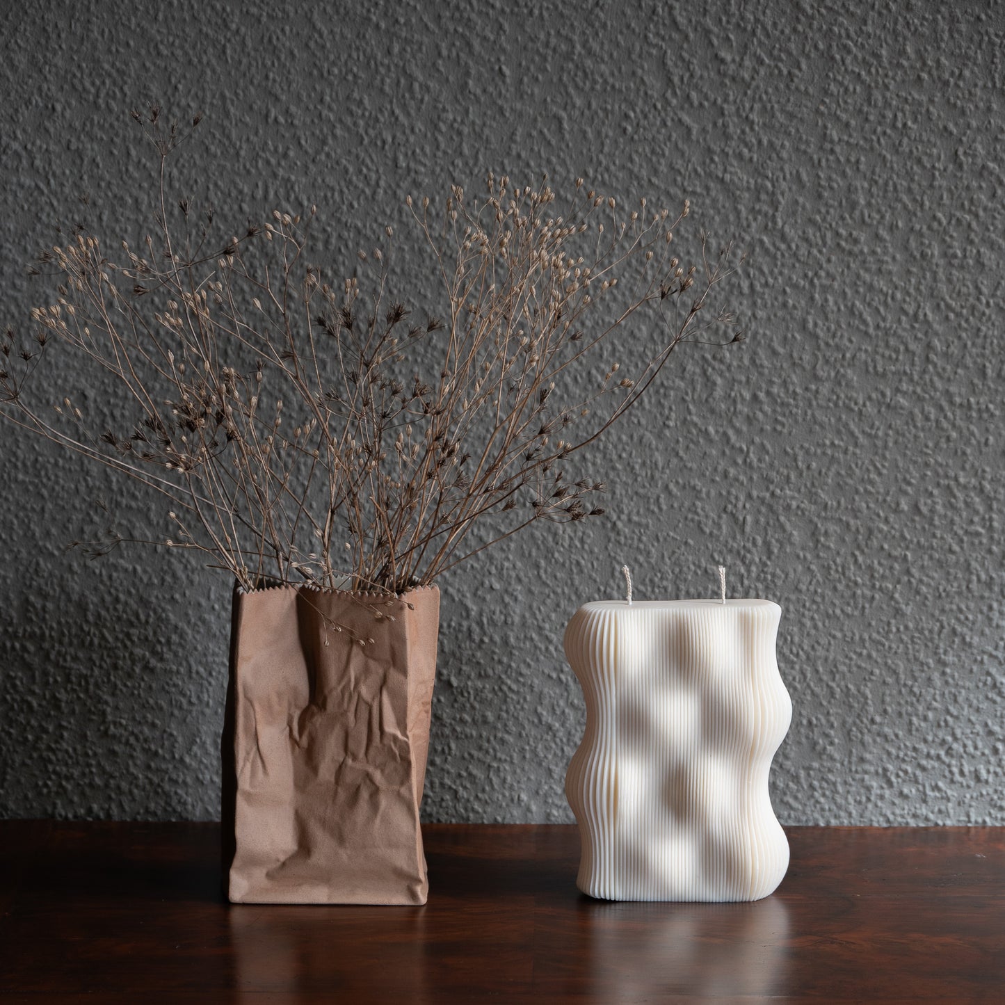 Væg Kerze in natürlichem Cremeton – Designkerzen aus Rapswachs, handgefertigt in Hamburg. 100 % pflanzlich & nachhaltig. Studio Z. Skulpturale Wellenform wie eine horizontale Wachswand. Dekoriert auf barocker Kommode neben brauner Tüten- Vase vor grauer Wand umwunden von Trockenblumen.