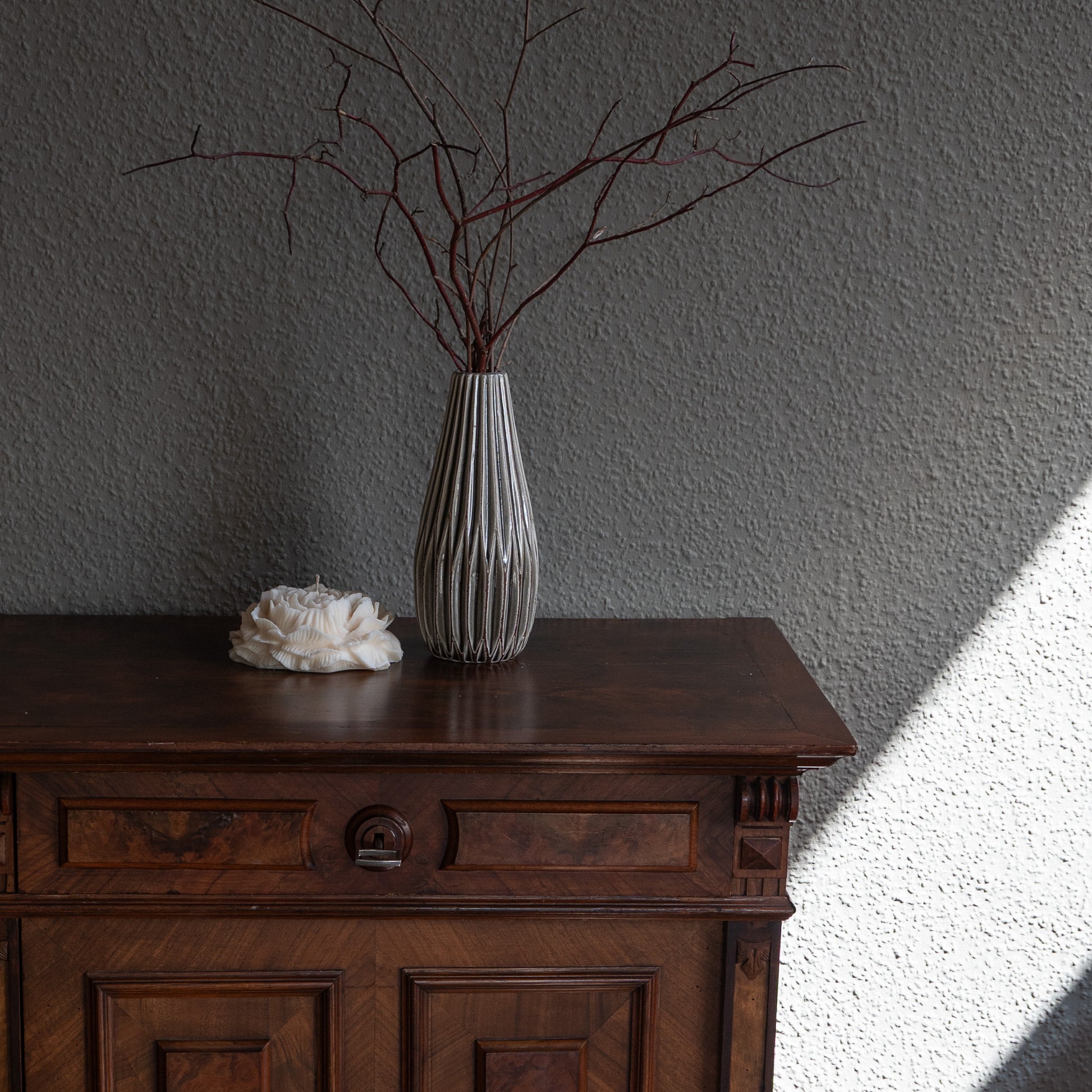 Kerze Bløma in großer Pfingstrosenform, große Blüte in natürlichem Cremeton, Designkerzen, Rapswachs, handgefertigt in Hamburg, 100 % pflanzlich & nachhaltig. Studio Z. Dekoriert auf barockem Tisch vor grauer Wand neben geriffelter hoher Vase in grau mit Ästen.