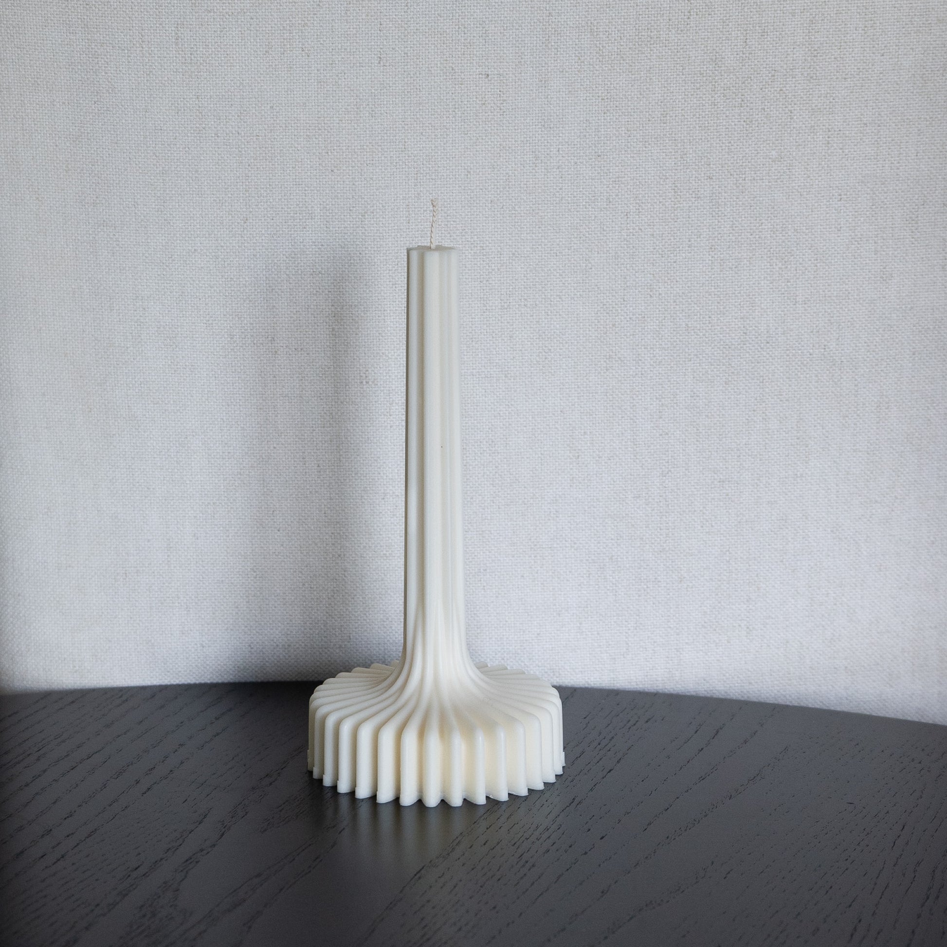 Kerze Fylda, plissierte Stabkerze in natürlichem Cremeton – skulpturale längliche Designkerzen, Rapswachs, handgefertigt in Hamburg, 100 % pflanzlich & nachhaltig. Studio Z. Geriffelte Struktur mit Plateaufuß, dekoriert auf schwarzem Tisch vor hellem Hintergrund.