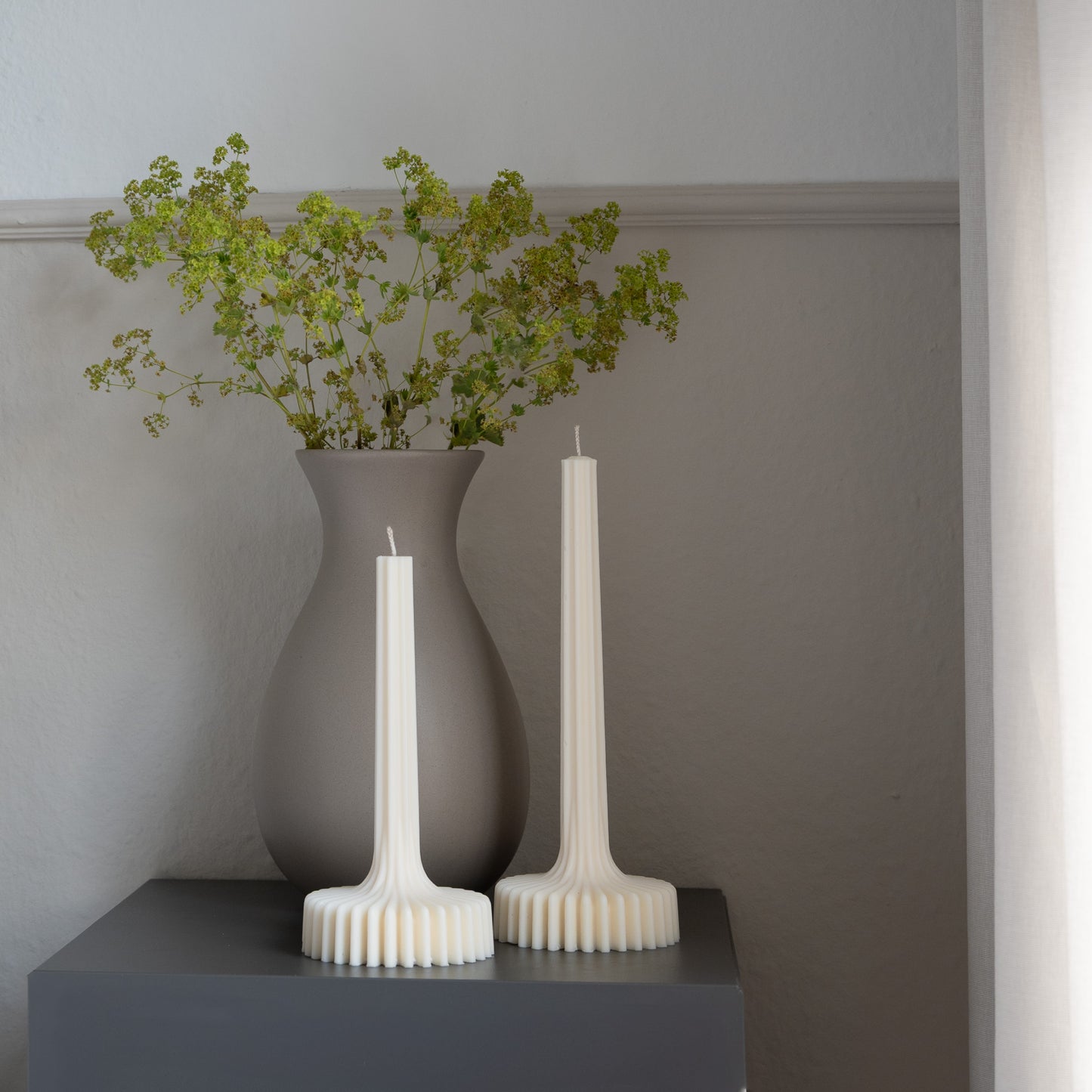Kerze Fylda, plissierte Stabkerze in natürlichem Cremeton – skulpturale längliche Designkerzen, Rapswachs, handgefertigt in Hamburg, 100 % pflanzlich & nachhaltig. Studio Z. Geriffelte Struktur mit Plateaufuß, dekoriert auf grauem Tisch neben Blumenvase.