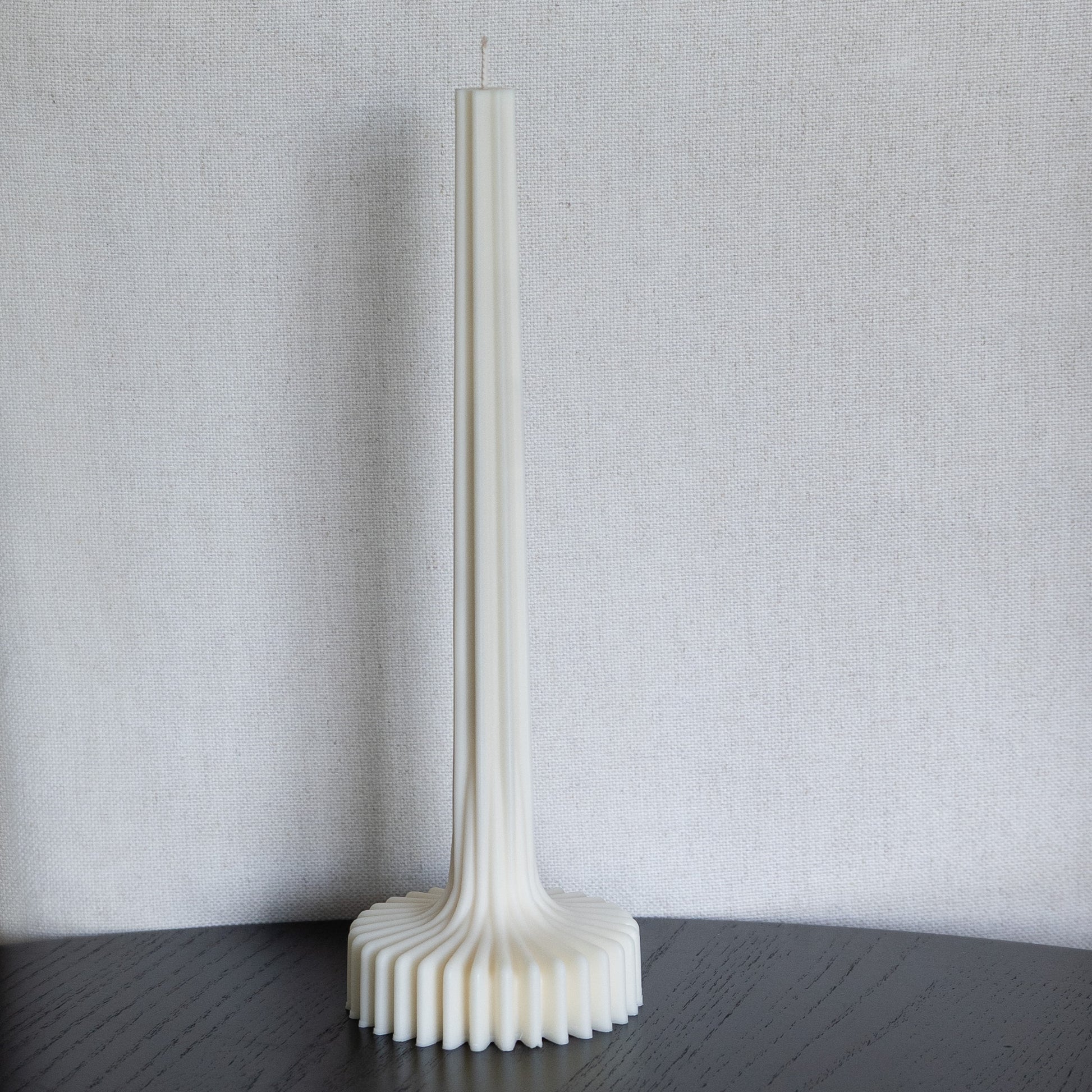 Kerze Fylda, plissierte Stabkerze in natürlichem Cremeton – skulpturale längliche Designkerzen, Rapswachs, handgefertigt in Hamburg, 100 % pflanzlich & nachhaltig. Studio Z. Geriffelte Struktur mit Plateaufuß, dekoriert auf schwarzem Tisch vor hellem Hintergrund.