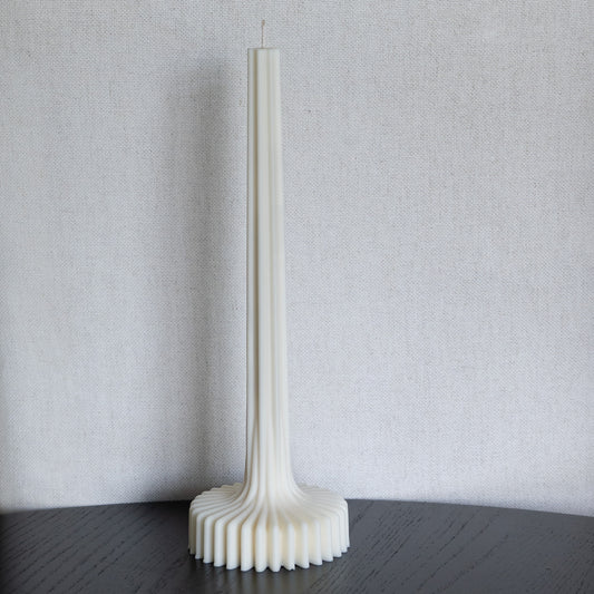 Kerze Fylda, plissierte Stabkerze in natürlichem Cremeton – skulpturale längliche Designkerzen, Rapswachs, handgefertigt in Hamburg, 100 % pflanzlich & nachhaltig. Studio Z. Geriffelte Struktur mit Plateaufuß, dekoriert auf schwarzem Tisch vor hellem Hintergrund.