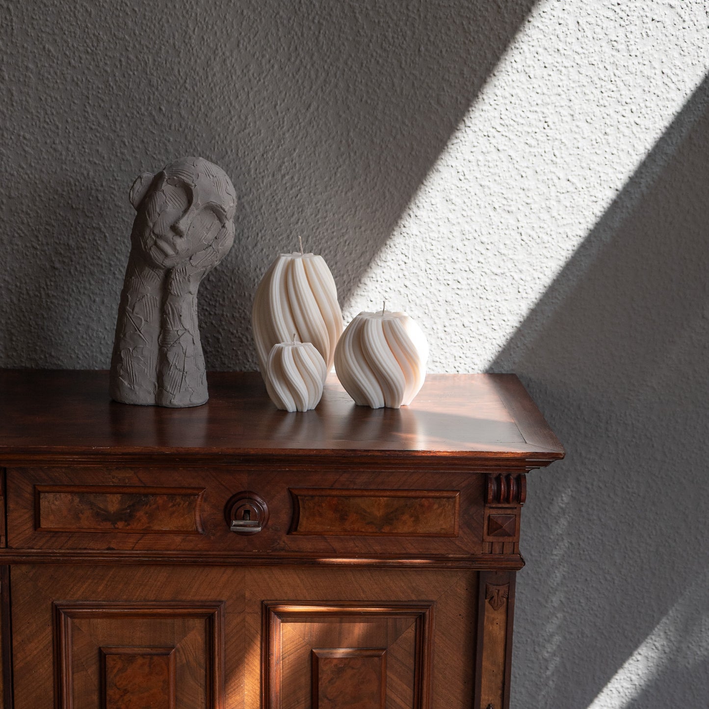 3er Kerzen Set Bøluna in natürlichem Cremeton – skulpturale Designkerzen, Rapswachs, handgefertigt in Hamburg, 100 % pflanzlich & nachhaltig. Studio Z. Stumpenkerzen in Wellenform, geschwungen und bauchig. Dekoriert auf barocker brauner Kommode im Licht der Sonne mit Schattenspielen neben nachdenklicher Betonfigur.