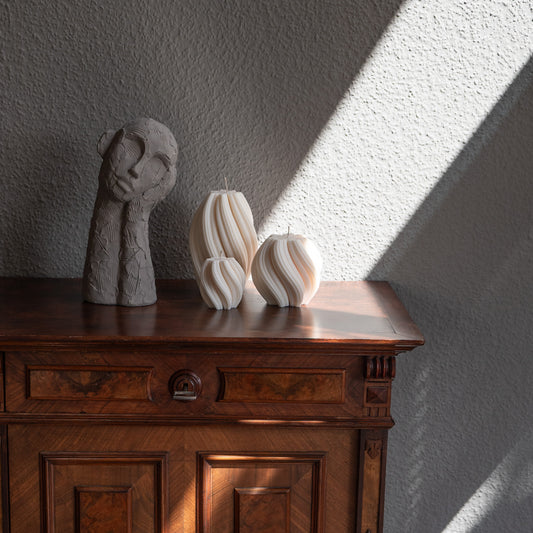 3er Kerzen Set Bøluna in natürlichem Cremeton – skulpturale Designkerzen, Rapswachs, handgefertigt in Hamburg, 100 % pflanzlich & nachhaltig. Studio Z. Stumpenkerzen in Wellenform, geschwungen und bauchig. Dekoriert auf barocker brauner Kommode im Licht der Sonne mit Schattenspielen neben nachdenklicher Betonfigur.