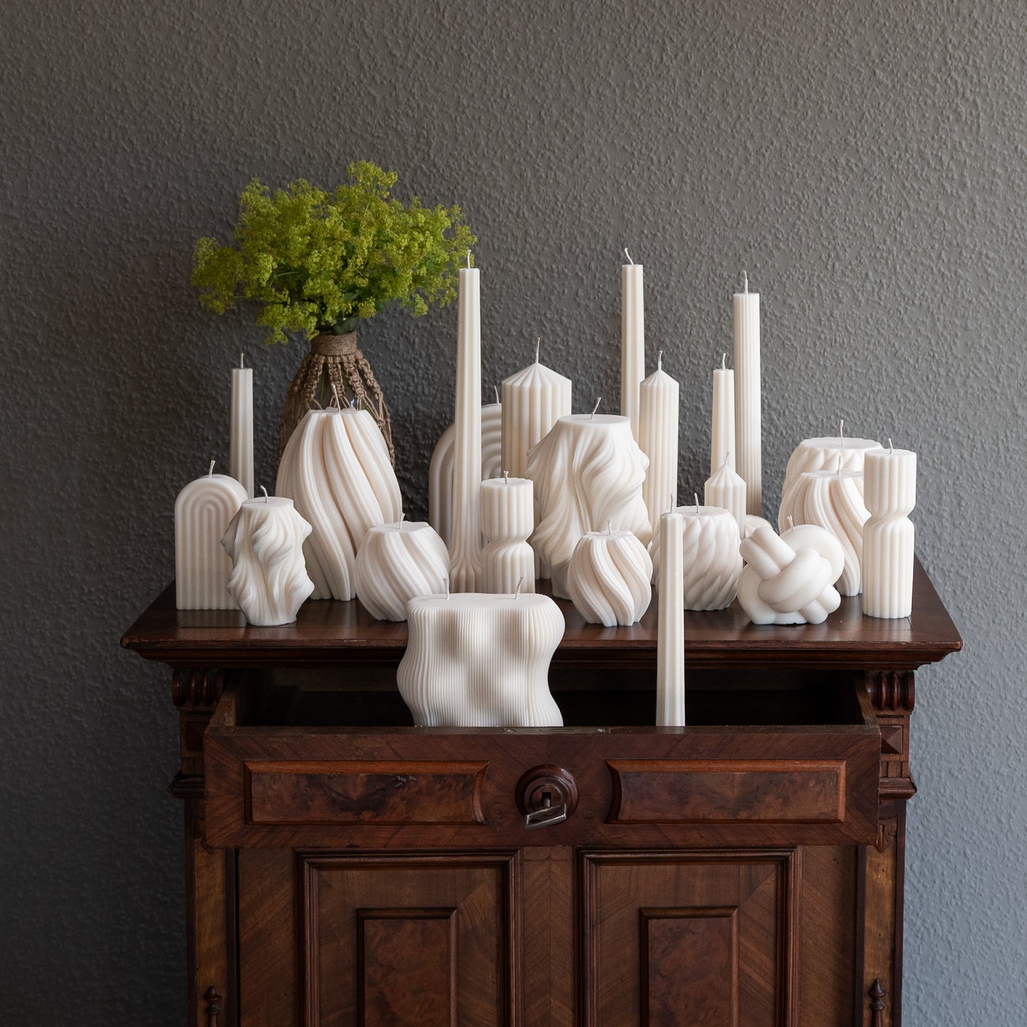 Auswahl  diverser Design-Kerze in natürlichem Cremeton  auf barocker Kommode vor grauer Wand neben Vase mit Ästen. Designkerzen aus Rapswachs, handgefertigt in Hamburg. 100 % pflanzlich & nachhaltig. Studio Z. Strøme Væg Lys Bøluna Slør Knøt Bølge Fylda 