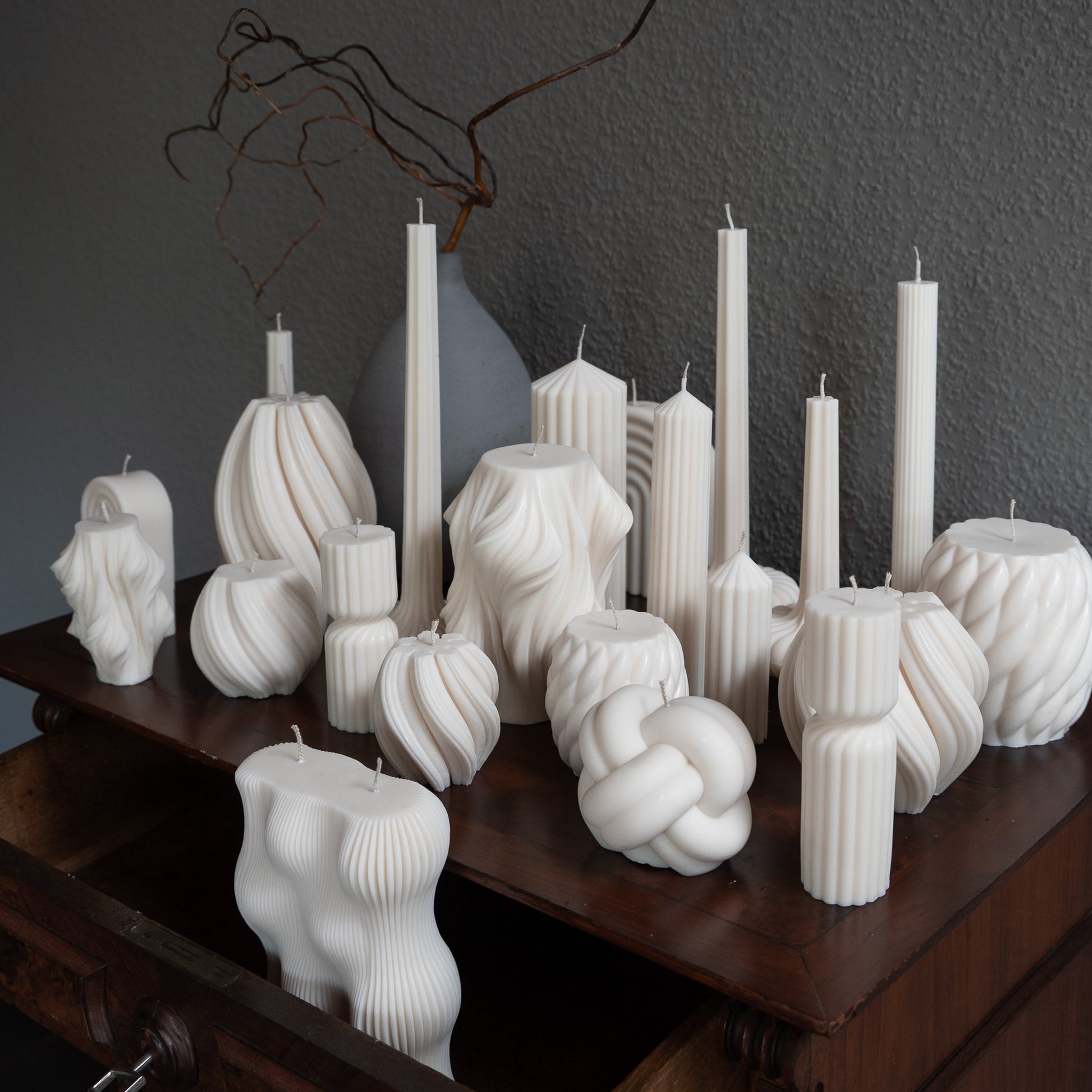 Auswahl  diverser Design-Kerze in natürlichem Cremeton  auf barocker Kommode vor grauer Wand neben Vase mit Ästen. Designkerzen aus Rapswachs, handgefertigt in Hamburg. 100 % pflanzlich & nachhaltig. Studio Z. Strøme Væg Lys Bøluna Slør Knøt Bølge Fylda 