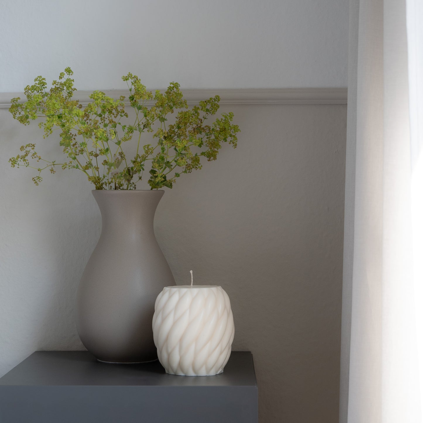 Knøv Kerze in natürlichem Cremeton – skulpturale Designkerzen, Rapswachs, handgefertigt in Hamburg, 100 % pflanzlich & nachhaltig. Studio Z. Wellenstruktur, bauchig, gewebt. Bauchige, gewebt wirkende Kerze dekoriert auf grauem Tisch neben Vase mit Blumen und Leinenvorhang.