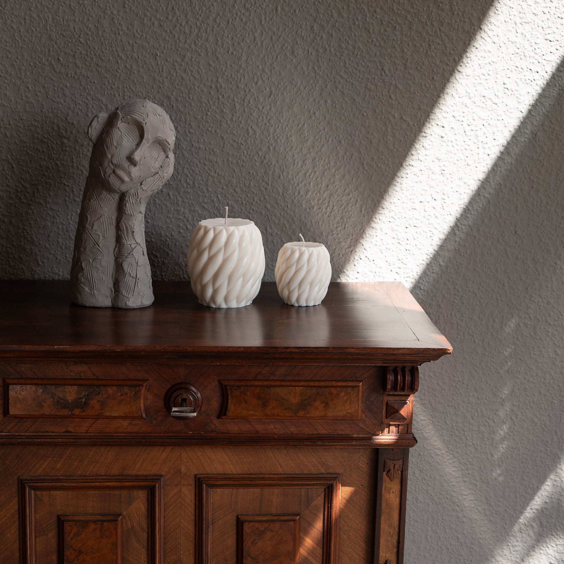 Zweier Kerzen-Set Knøv in natürlichem Cremeton – skulpturale Designkerzen, Rapswachs, handgefertigt in Hamburg, 100 % pflanzlich & nachhaltig. Studio Z. Wellenstruktur, bauchig, gewebt. Dekoriert in zwei Größen auf barocker Kommode im Schatten-Lichtspiel der Sonne neben nachdenklicher Betonfigur.