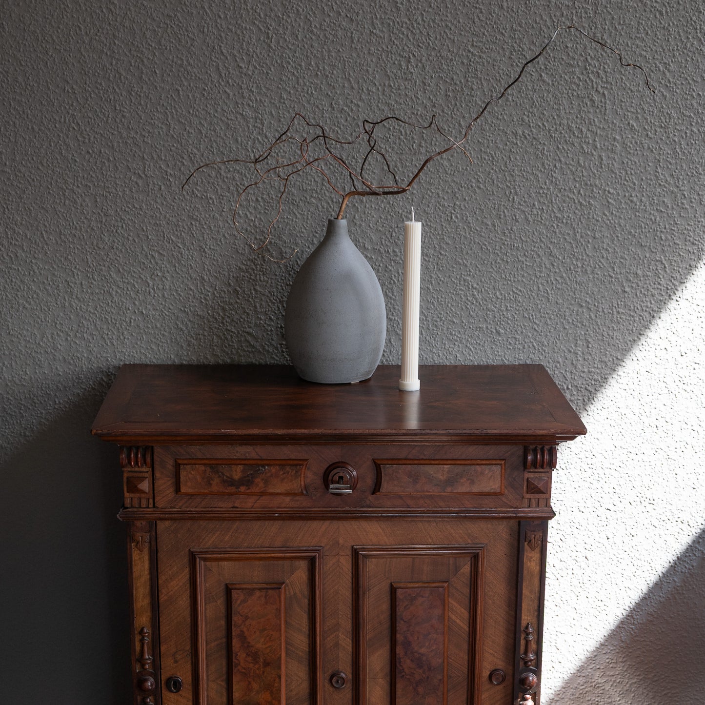 Kerze Lys, geradlininge Stumpenkerze in natürlichem Cremeton – geriffelte längliche Designkerzen, Rapswachs, handgefertigt in Hamburg, 100 % pflanzlich & nachhaltig. Studio Z. Dekoriert auf barocker Kommode vor grauem  Hintergrund neben geriffelter Vase mit Ästen.