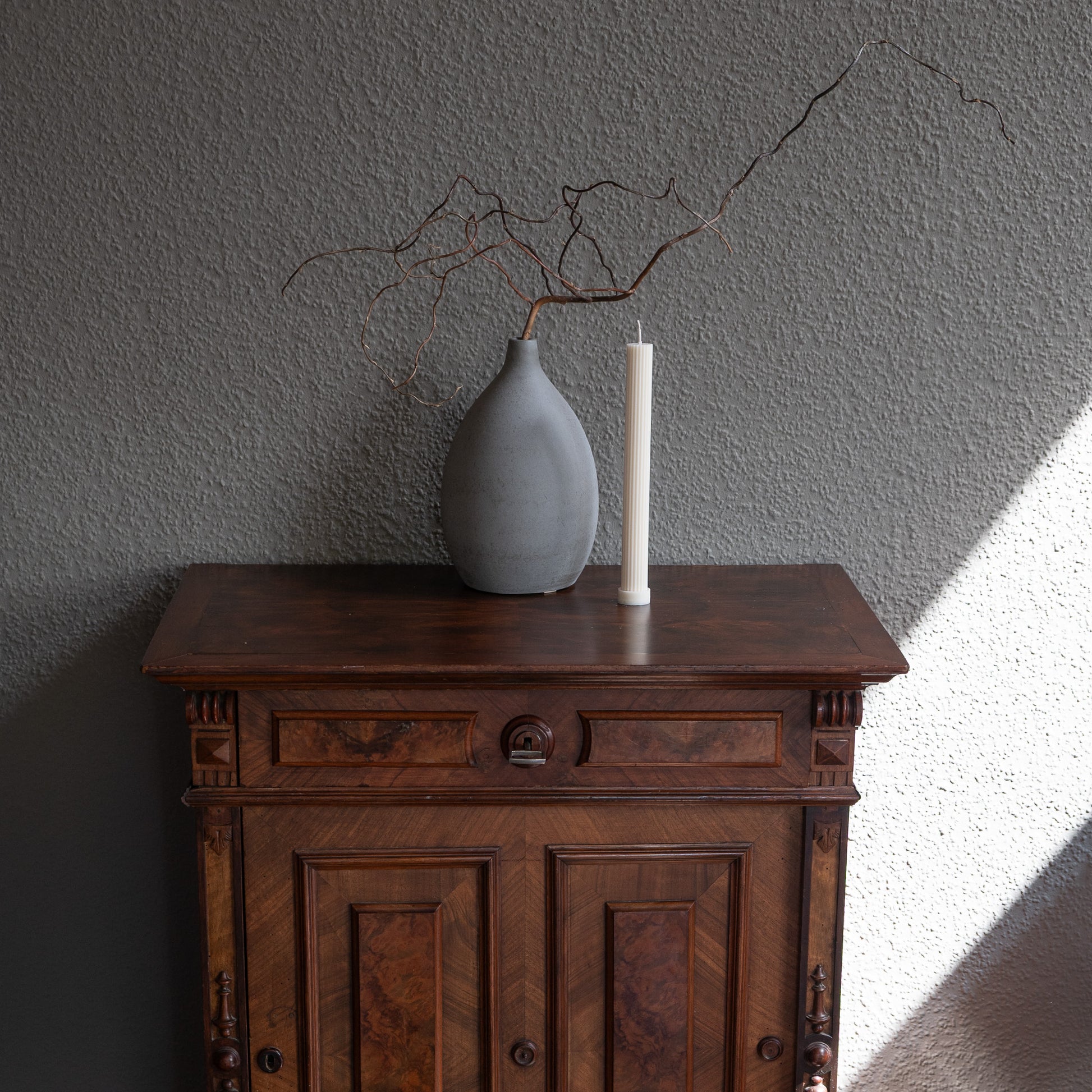 Kerze Lys, geradlininge Stumpenkerze in natürlichem Cremeton – geriffelte längliche Designkerzen, Rapswachs, handgefertigt in Hamburg, 100 % pflanzlich & nachhaltig. Studio Z. Dekoriert auf barocker Kommode vor grauem  Hintergrund neben geriffelter Vase mit Ästen.