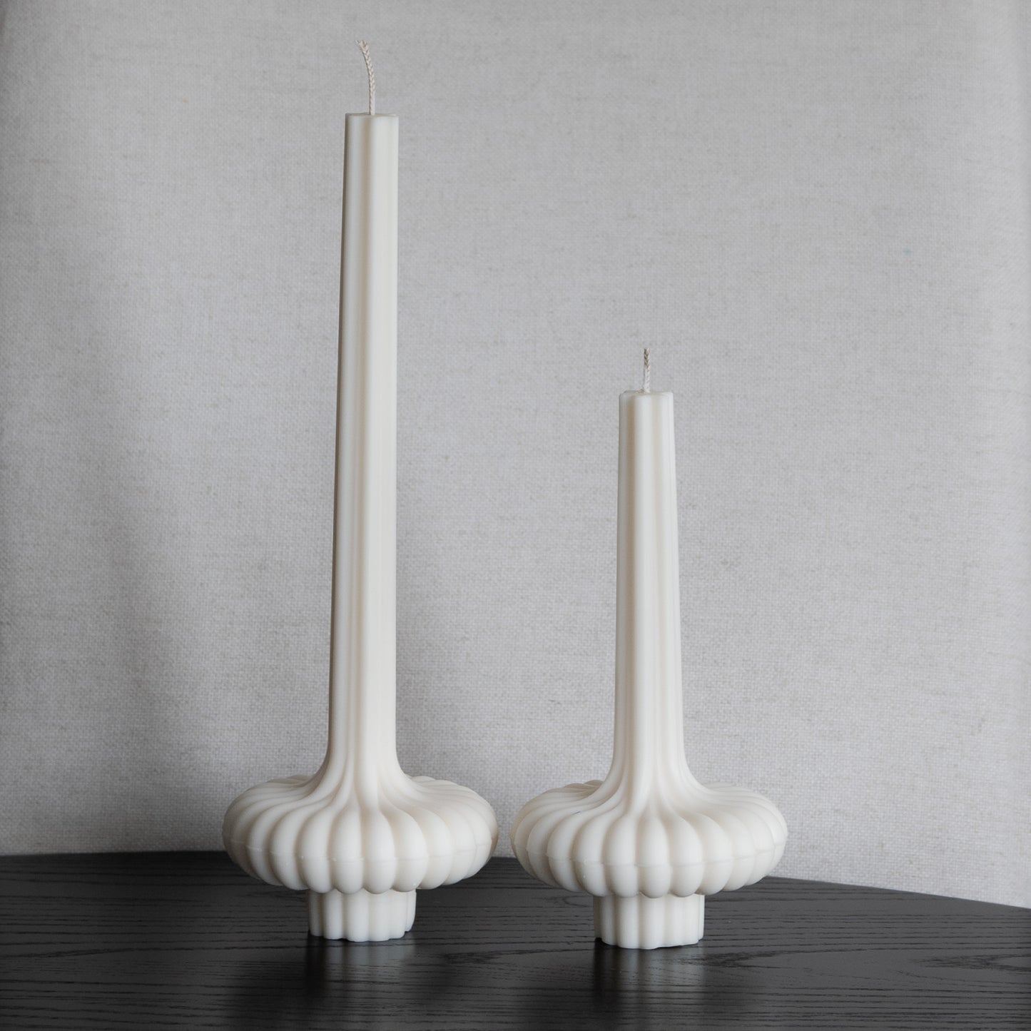 Zweier Set Skæva Kerze in natürlichem Cremeton – skulpturale Designkerzen aus Rapswachs, handgefertigt in Hamburg, 100 % pflanzlich & nachhaltig. Studio Z. Mit plissierter Struktur, markantem Fuß und skulpturaler Form. Dekoriert nebeneinander in zwei Größen auf schwarzem Tisch vor heller Leinenstruktur.