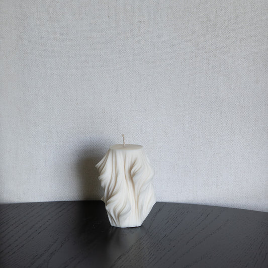 Skulpturale Design Kerze in Naturfarbe. Handgefertigt in Hamburg – Statement-Dekor im dänischen Design. 100 % natürliche, nachhaltige Materialien. Schleier Optik. Dekoriert auf schwarzem Tisch vor Leinen Stuhl.