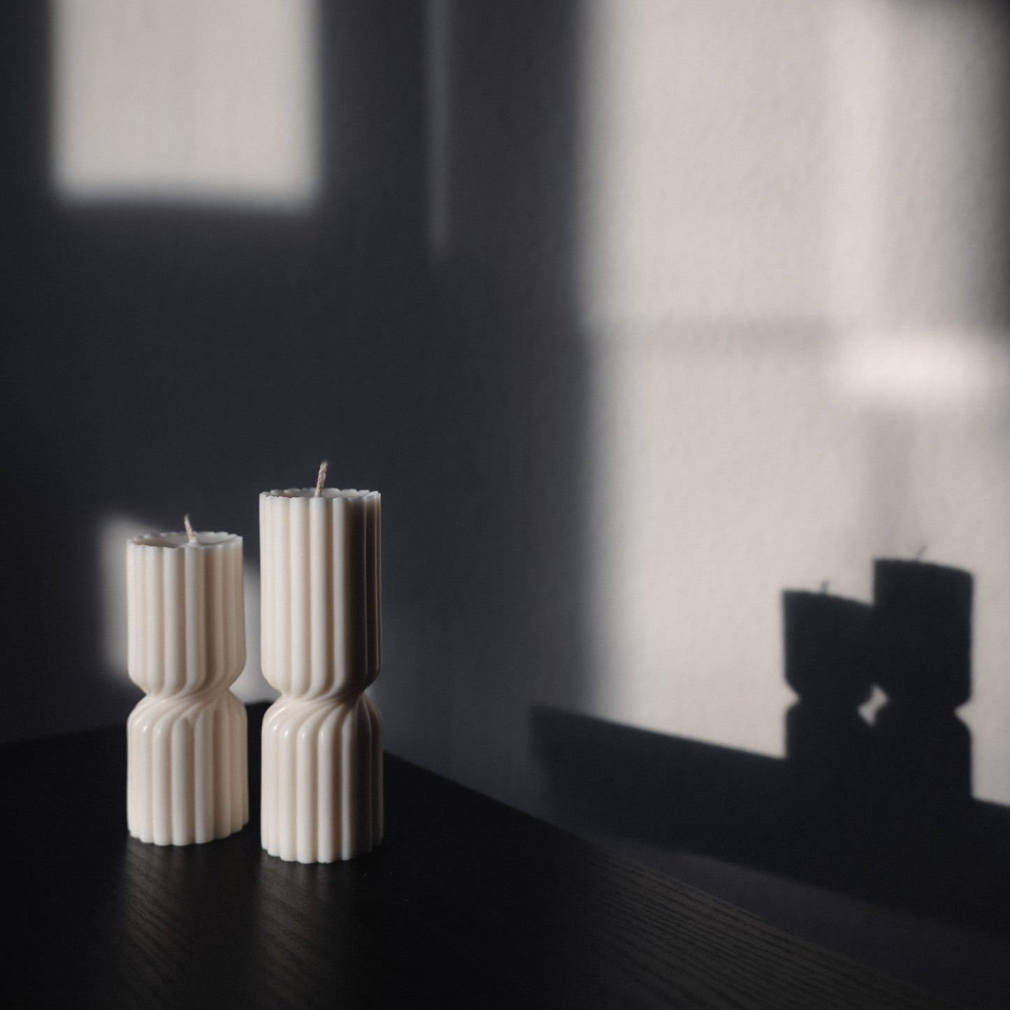 Zweier Kerzen-Set Strøme, mittig gedrehte Stumpenkerze in natürlichem Cremeton. Skulpturale längliche Designkerzen, Rapswachs, handgefertigt in Hamburg, 100 % pflanzlich & nachhaltig. Studio Z. Geriffelte Struktur, dekoriert auf schwarzem Tisch mit rückwärts im Schatten abgebildetem Spiegelbild der Formen.
