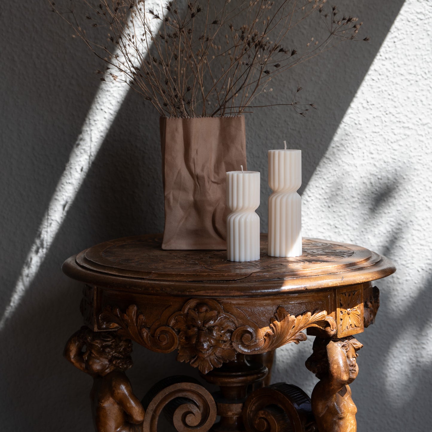 Kerze Strøme, gedrehte Stumpenkerze in natürlichem Cremeton – skulpturale längliche Designkerzen, Rapswachs, handgefertigt in Hamburg, 100 % pflanzlich & nachhaltig. Studio Z. Geriffelte Struktur., mittig gedreht Dekoriert auf barockem Tisch vneben Vase mit trockenblumen und Sonnenlicht und Schatten seitlich.