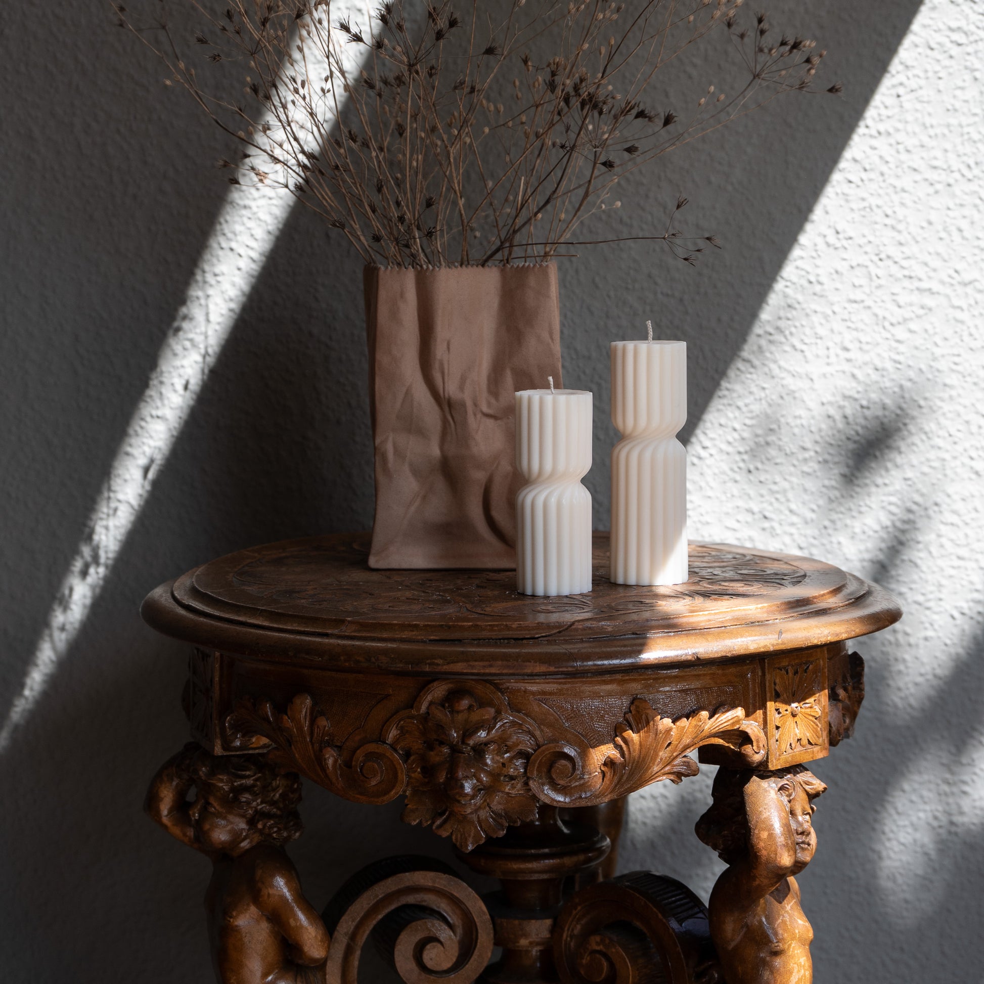 Kerze Strøme, gedrehte Stumpenkerze in natürlichem Cremeton – skulpturale längliche Designkerzen, Rapswachs, handgefertigt in Hamburg, 100 % pflanzlich & nachhaltig. Studio Z. Geriffelte Struktur., mittig gedreht Dekoriert auf barockem Tisch vneben Vase mit trockenblumen und Sonnenlicht und Schatten seitlich.