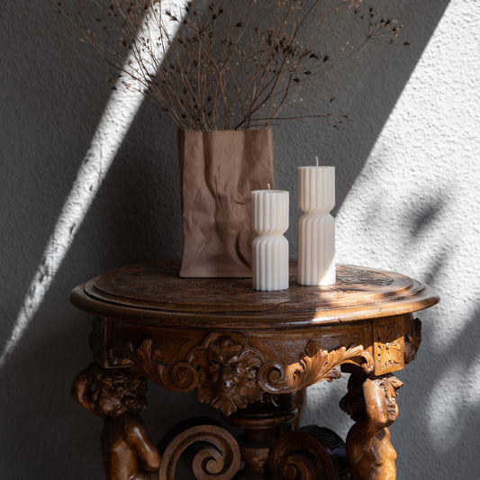 Kerze Strøme, gedrehte Stumpenkerze in natürlichem Cremeton – skulpturale längliche Designkerzen, Rapswachs, handgefertigt in Hamburg, 100 % pflanzlich & nachhaltig. Studio Z. Geriffelte Struktur., mittig gedreht Dekoriert auf barockem Tisch vneben Vase mit trockenblumen und Sonnenlicht und Schatten seitlich.