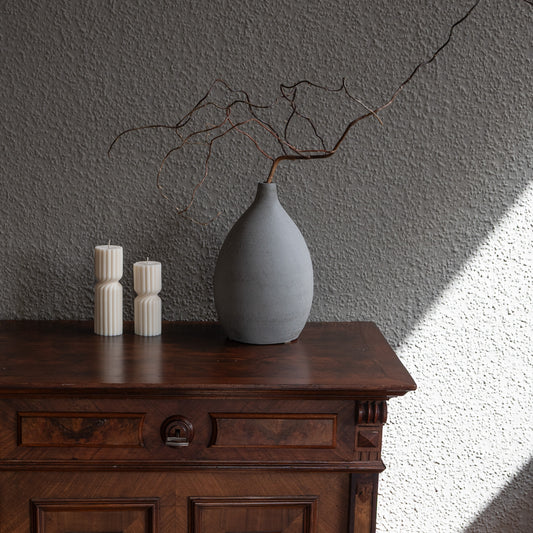 Kerze Strøme gedrehte Stumpenkerze in natürlichem Cremeton – skulpturale längliche Designkerzen, Rapswachs, handgefertigt in Hamburg, 100 % pflanzlich & nachhaltig. Studio Z. Geriffelte Struktur., mittig gedreht. Dekoriert auf barockem Tisch neben grauer Vase und dunkler Wand und trocknen Ästen.