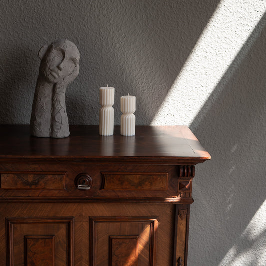 Zweier Kerzen-Set Strøme, mittig gedrehte Stumpenkerze in natürlichem Cremeton. Skulpturale längliche Designkerzen, Rapswachs, handgefertigt in Hamburg, 100 % pflanzlich & nachhaltig. Studio Z. Geriffelte Struktur, dekoriert auf barockem Tisch neben nachdenklicher Betonfigur und einfallendem Sonnenlicht und Schattenspiel.